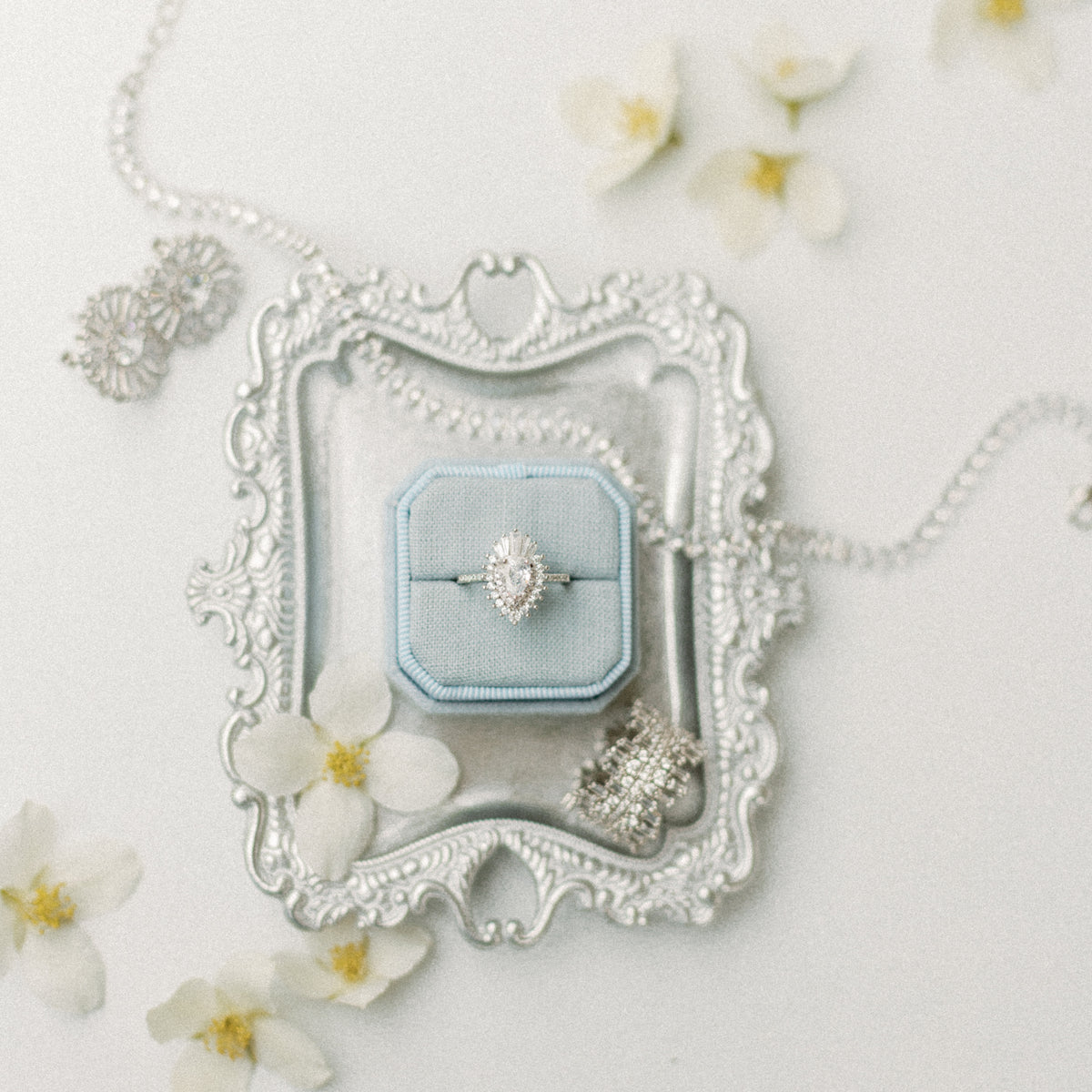 Ornate silver-tone styling tray with a light blue ring box holding a halo ring, styled with white blossoms
