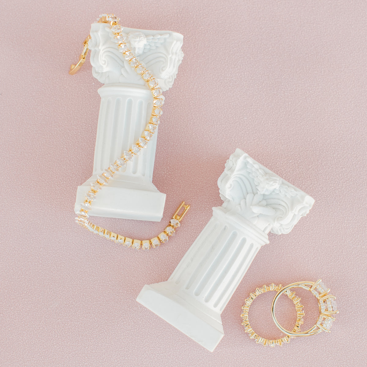 Mini white classical column pillars on blush, one draped with a gold-tone crystal bracelet, rings beside