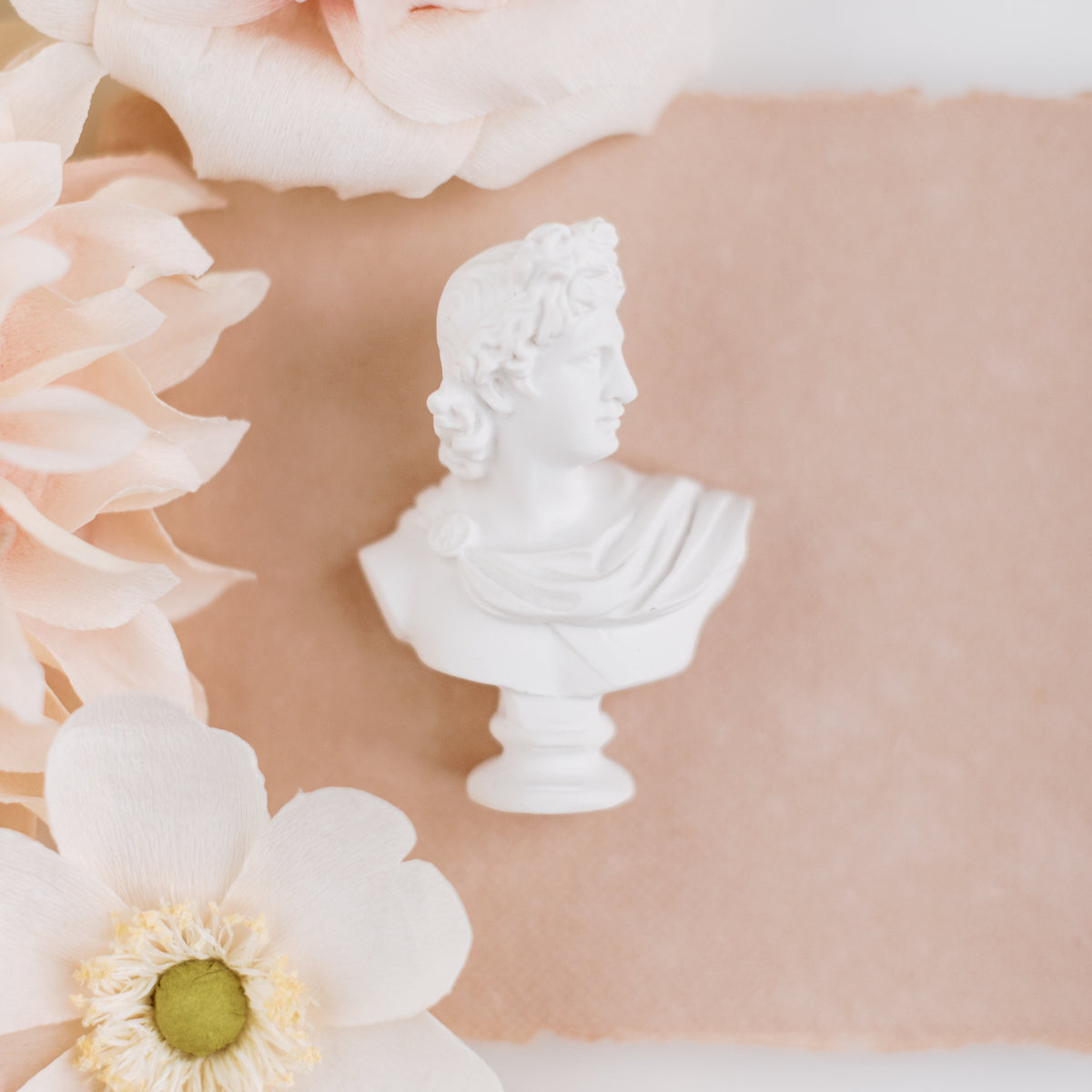 Mini white classical bust statue, three-quarter view on blush paper framed by peach and white flowers