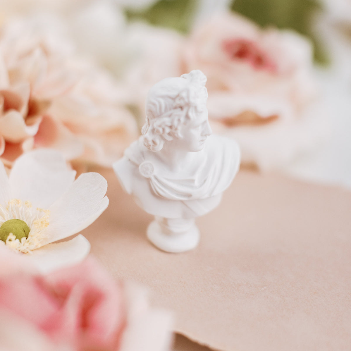 Mini white bust statue at eye level on blush paper, soft pink florals blurred behind