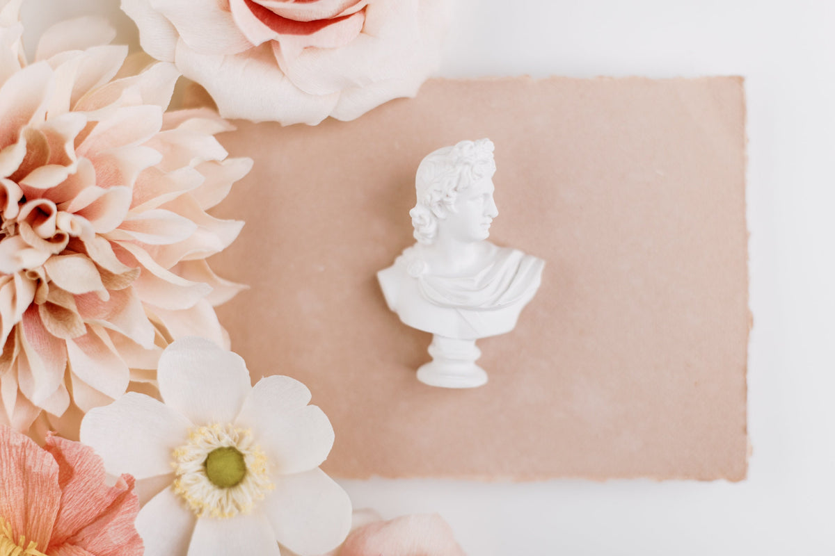 Mini white bust statue centered on a torn-edge blush paper, dahlias and roses along the left side
