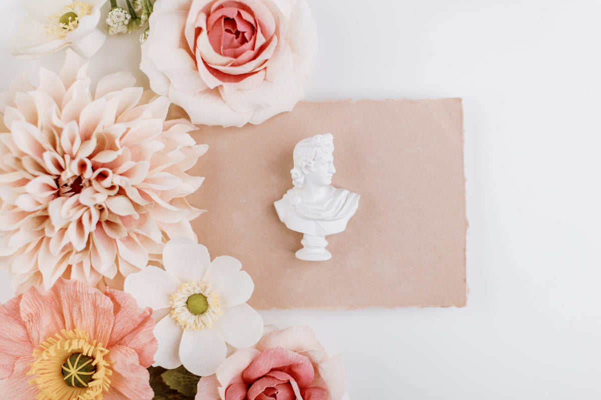 Mini white bust statue on a blush paper swatch beside pink and peach flowers