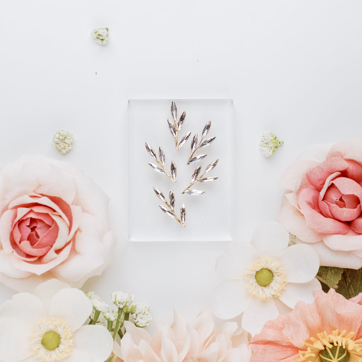 Gold-tone clear gem leaf sprigs arranged on a clear acrylic block, framed by blush roses and white blooms