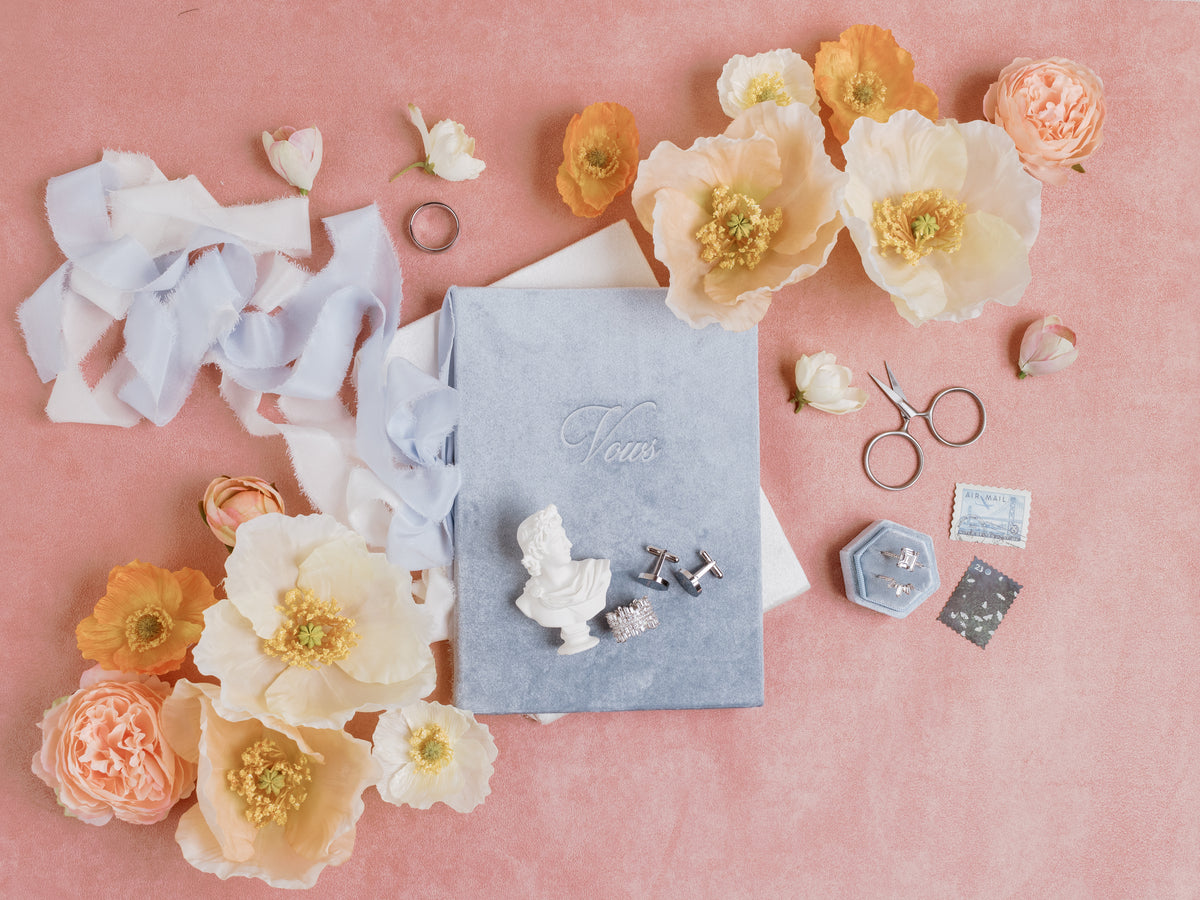Pink velvet flat lay with dusty blue invitation, mini white bust statue, ring box, scissors and poppies
