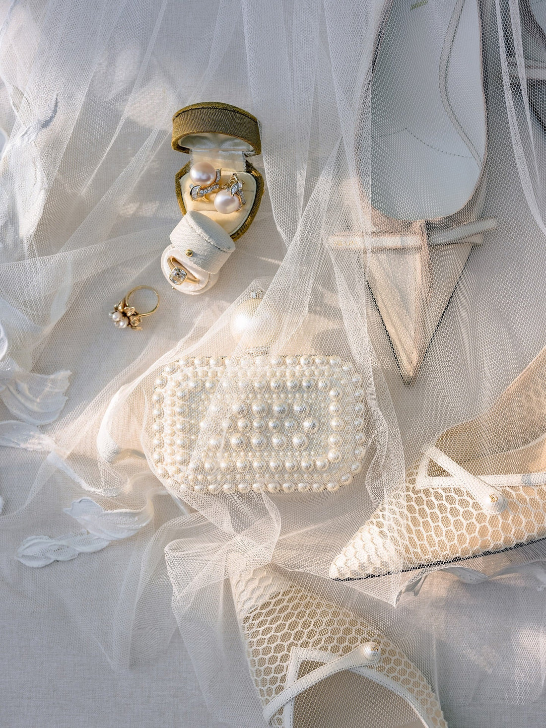 Bridal flat lay on tulle with pearl earrings in a sage heart ring box, the engagement ring in a Dove linen Georgian Oval ring box, a pearl clutch and mesh pumps