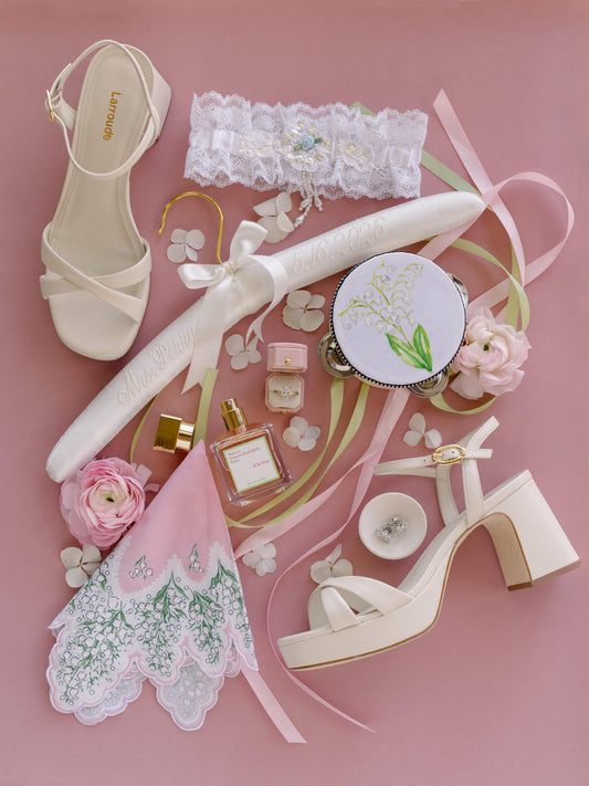 Bridal details flat lay with ivory sandals, lace garter, monogrammed hanger, perfume, lily of the valley handkerchief and the Radiant Roseate ring box