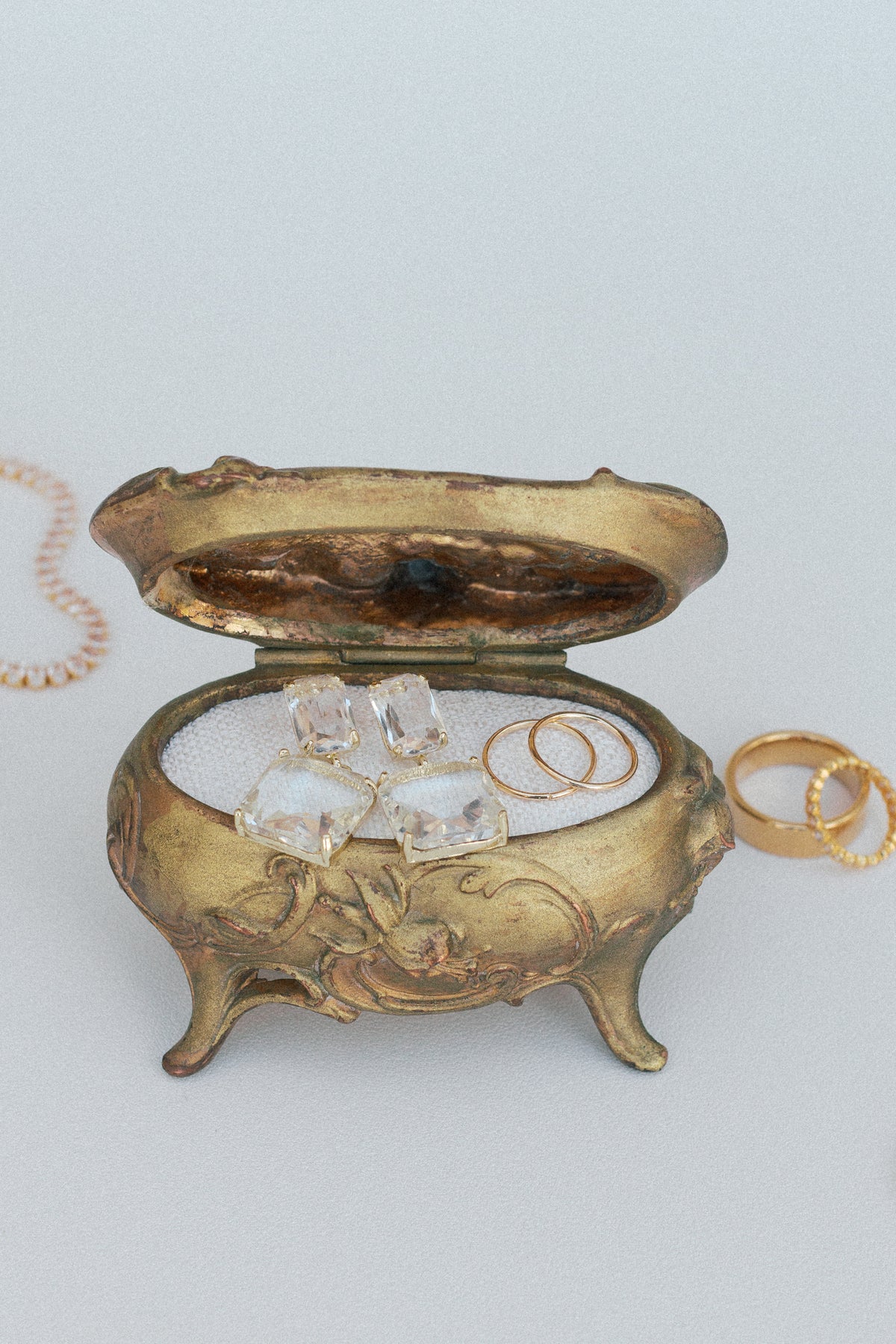 Gold-tone scrollwork footed box open with clear crystal earrings and gold rings on white lining