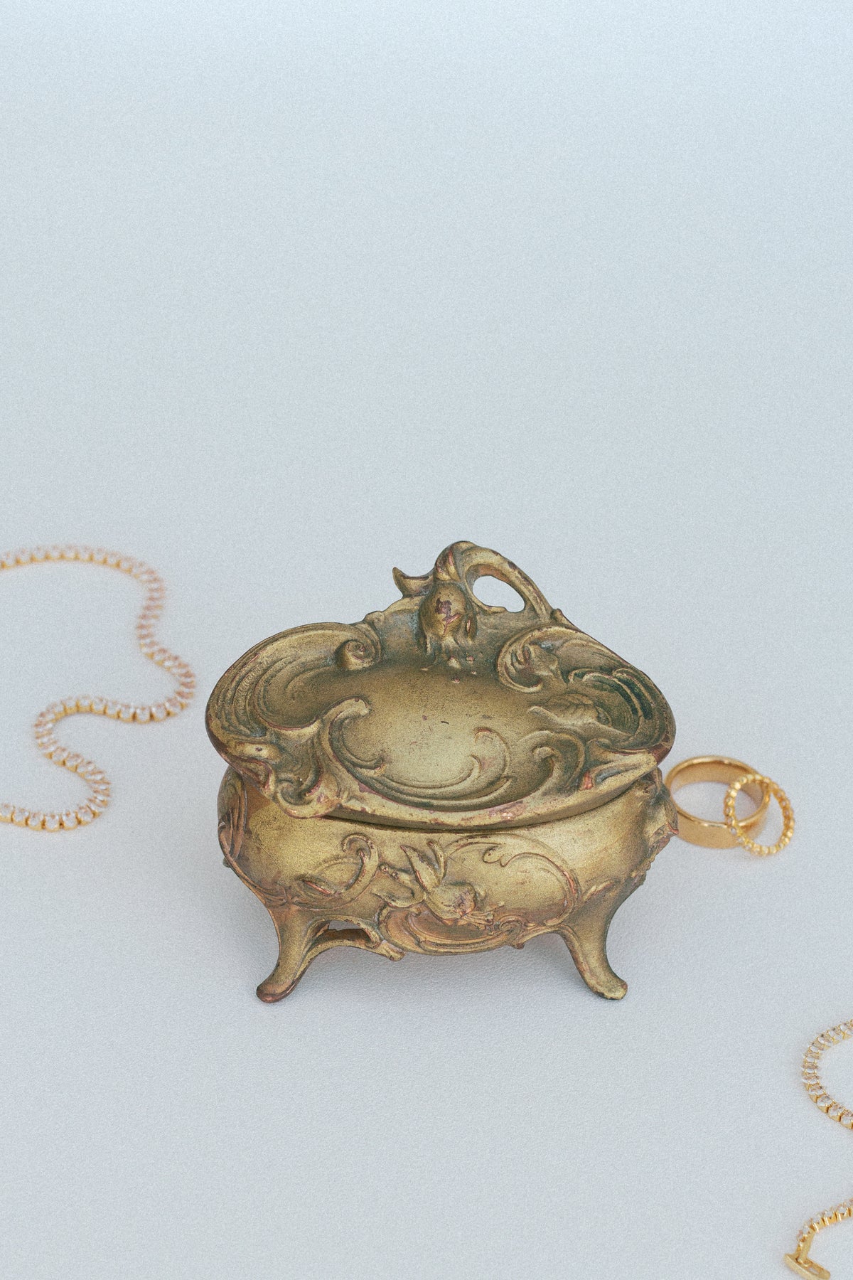 Gold-tone ornate scrollwork footed box closed, side view beside a gold chain and ring