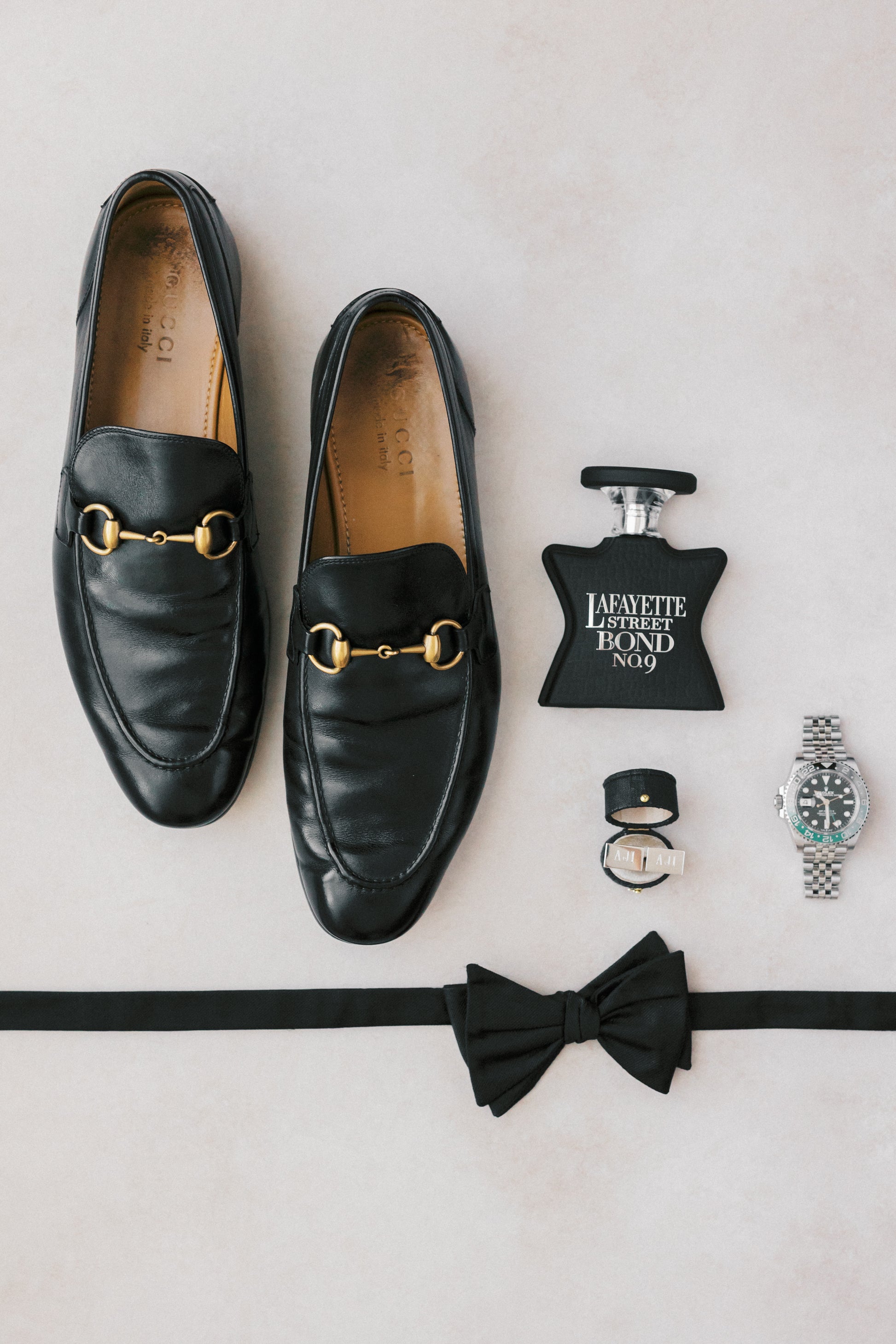 Black loafers with gold buckles, a black bow tie, a watch, and a bottle of cologne on a light gray background featuring the georgian oval ring box in black and raven